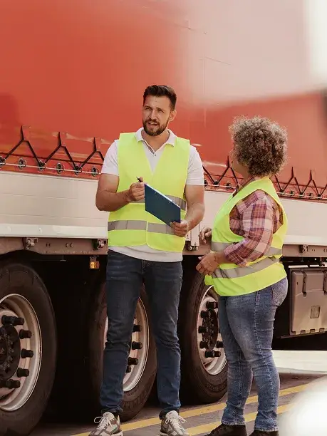 Two HGV drivers in hi-vis vests talking beside a red truck, one holding a clipboard